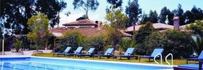 Zona de piscina con sillones de descanso y un edificio con tejado de azulejos.