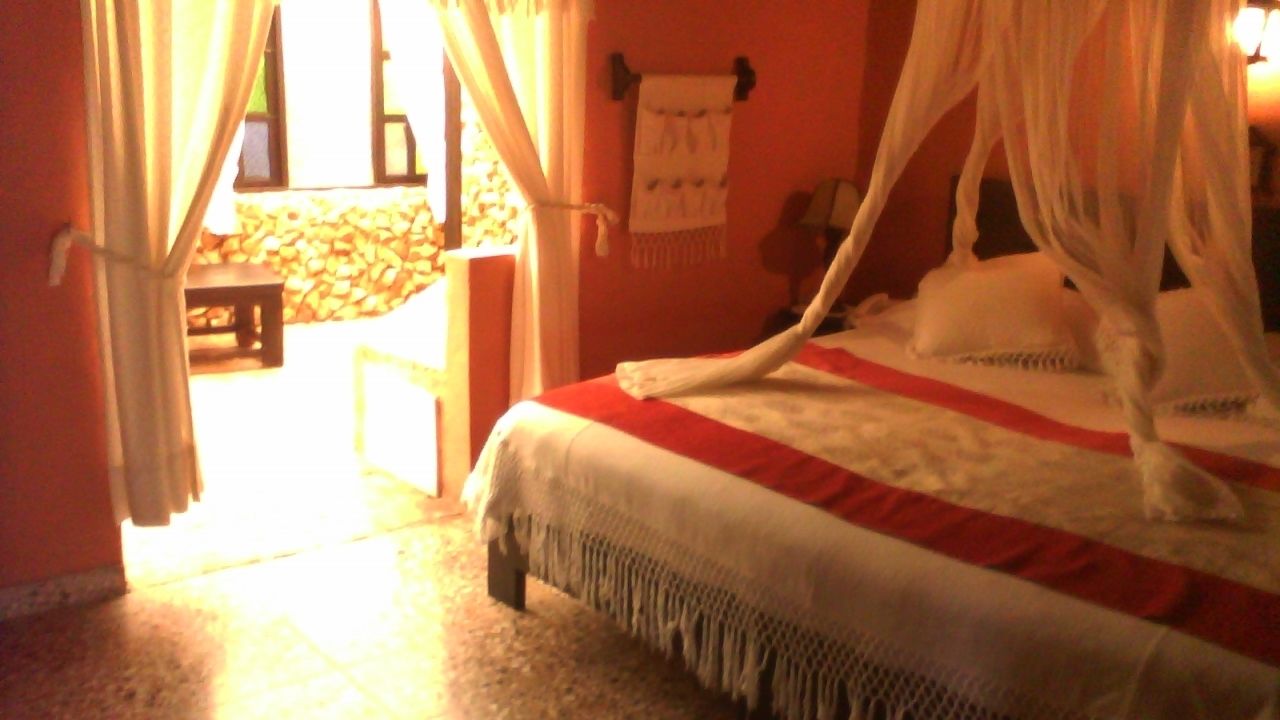 Una cama con dosel, una mesita pequeña y una silla en una habitación con pared de piedra y ventana.