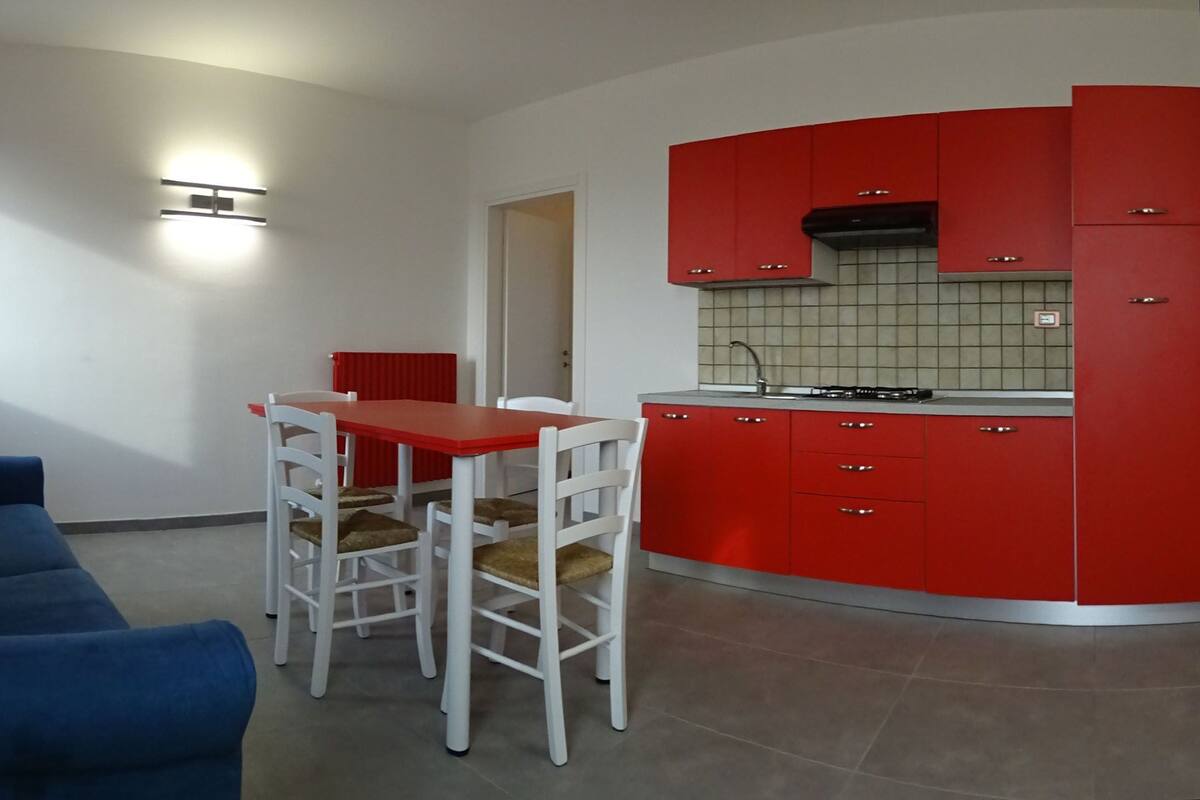 Um espaço de estar compacto com cozinha americana, área de refeições com mesa vermelha e cadeiras brancas, e sofá azul.