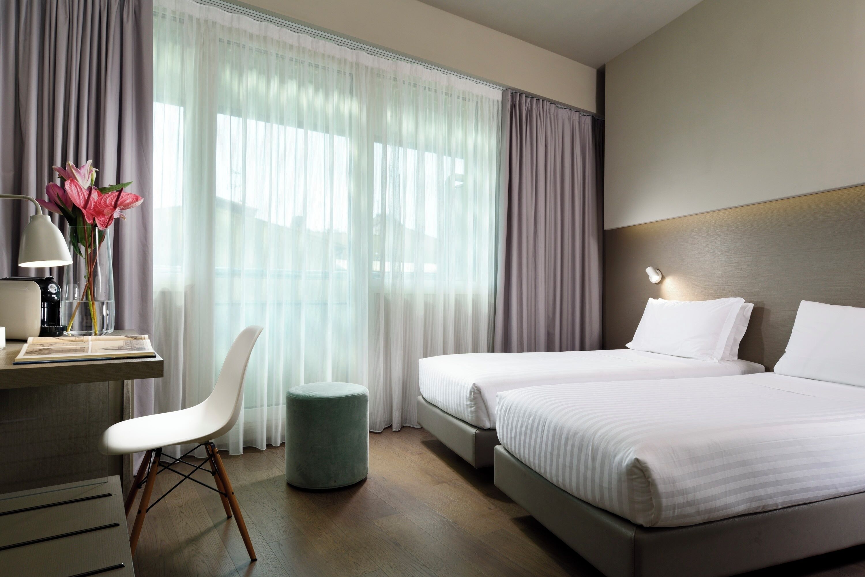 A hotel room with two beds, a desk with a lamp, a chair, and a small ottoman.