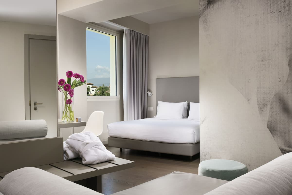 Een moderne hotelkamer met een bed, een zithoek met een bankje, een tafeltje en een raam met uitzicht op de stad.
