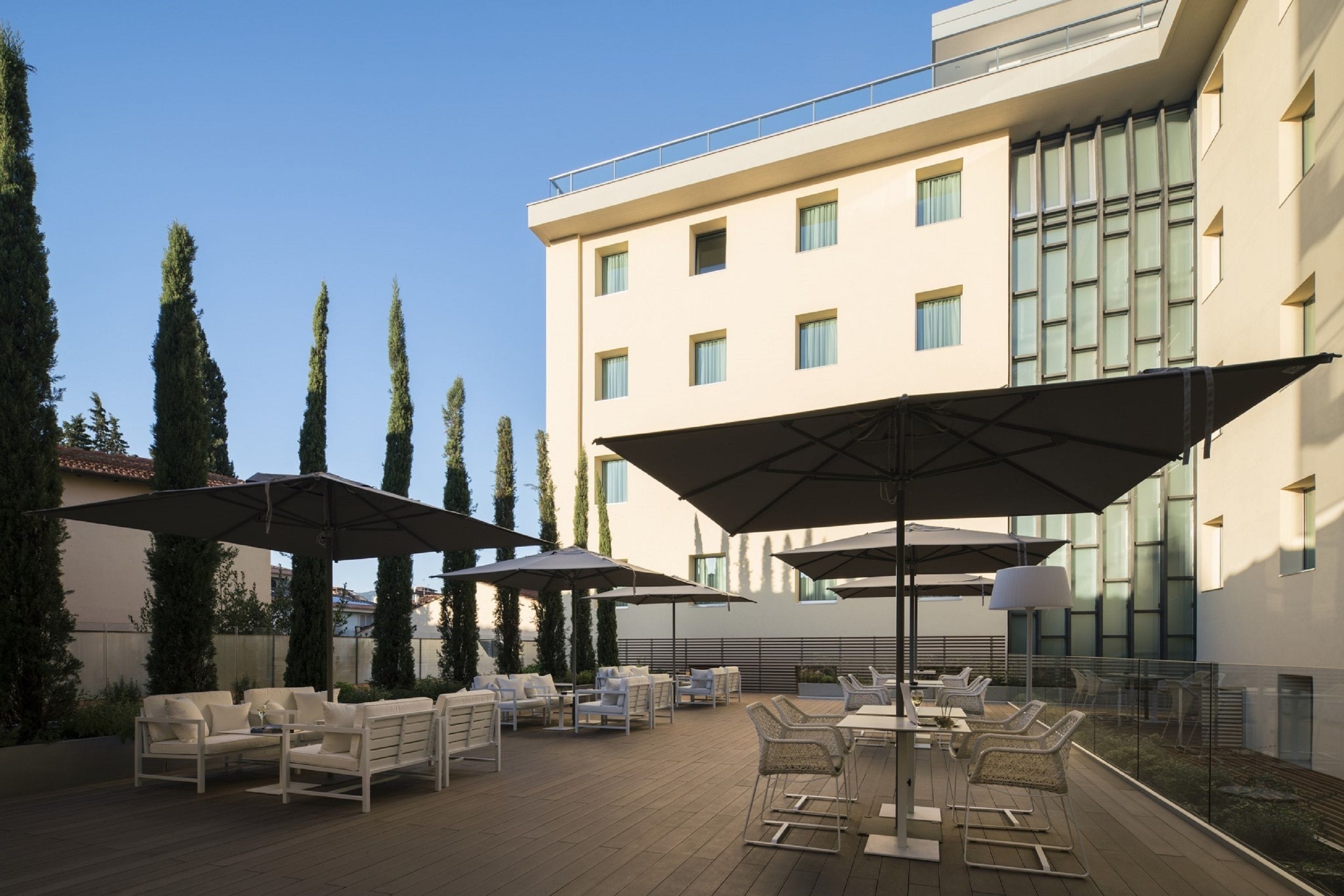 An outdoor patio with white furniture, large umbrellas, and a modern building in the background.