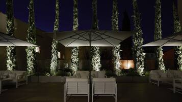 A patio area with white outdoor furniture and umbrellas, surrounded by tall, illuminated trees.