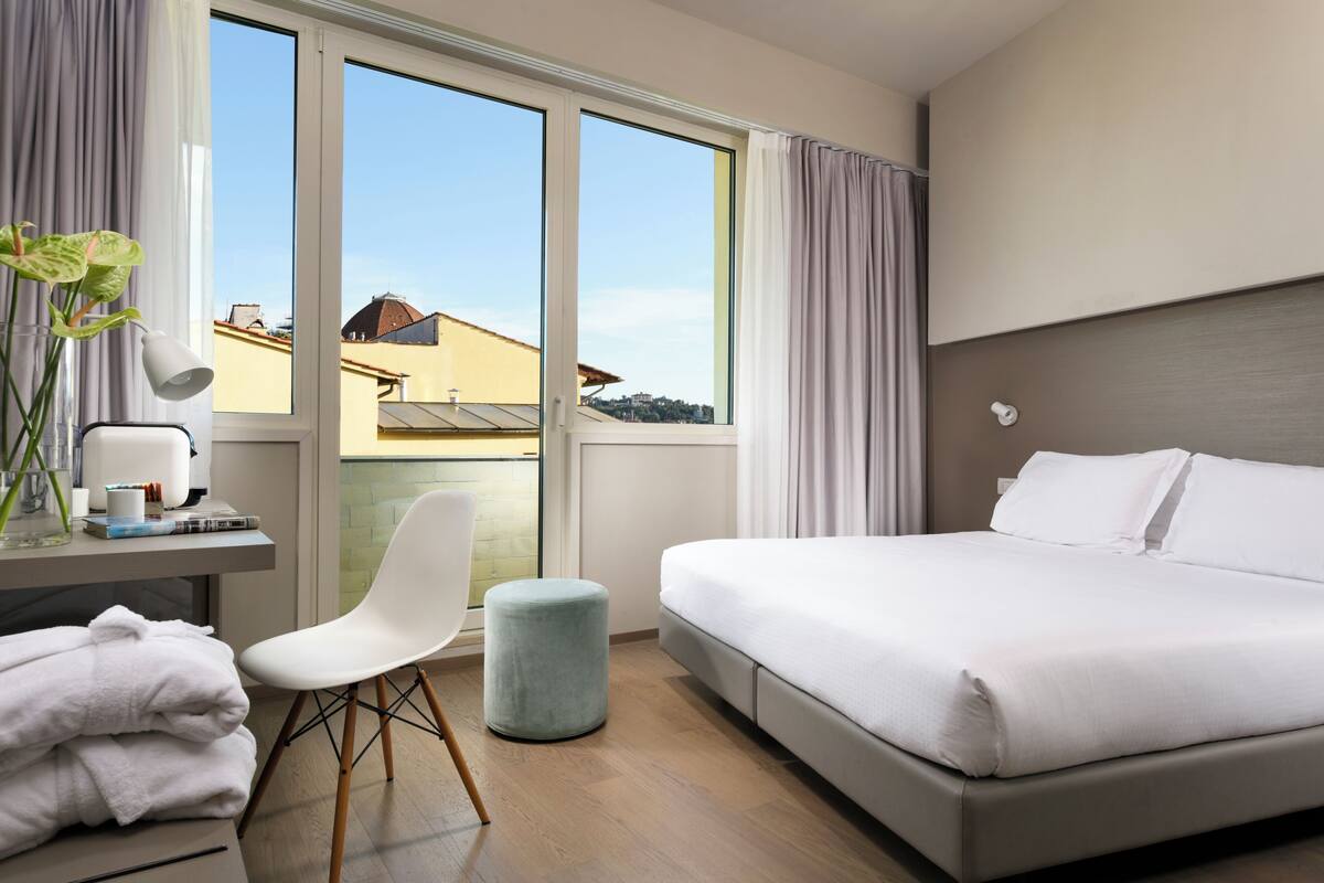 Een hotelkamer met een bed, een bureau met een lamp, een stoel en uitzicht op gebouwen door grote ramen.