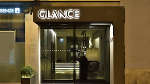 Ingången till ett hotell som heter GLANCE, med en modern design, inklusive en glasdörr och en synlig interiör med en lampa och en sittgrupp.