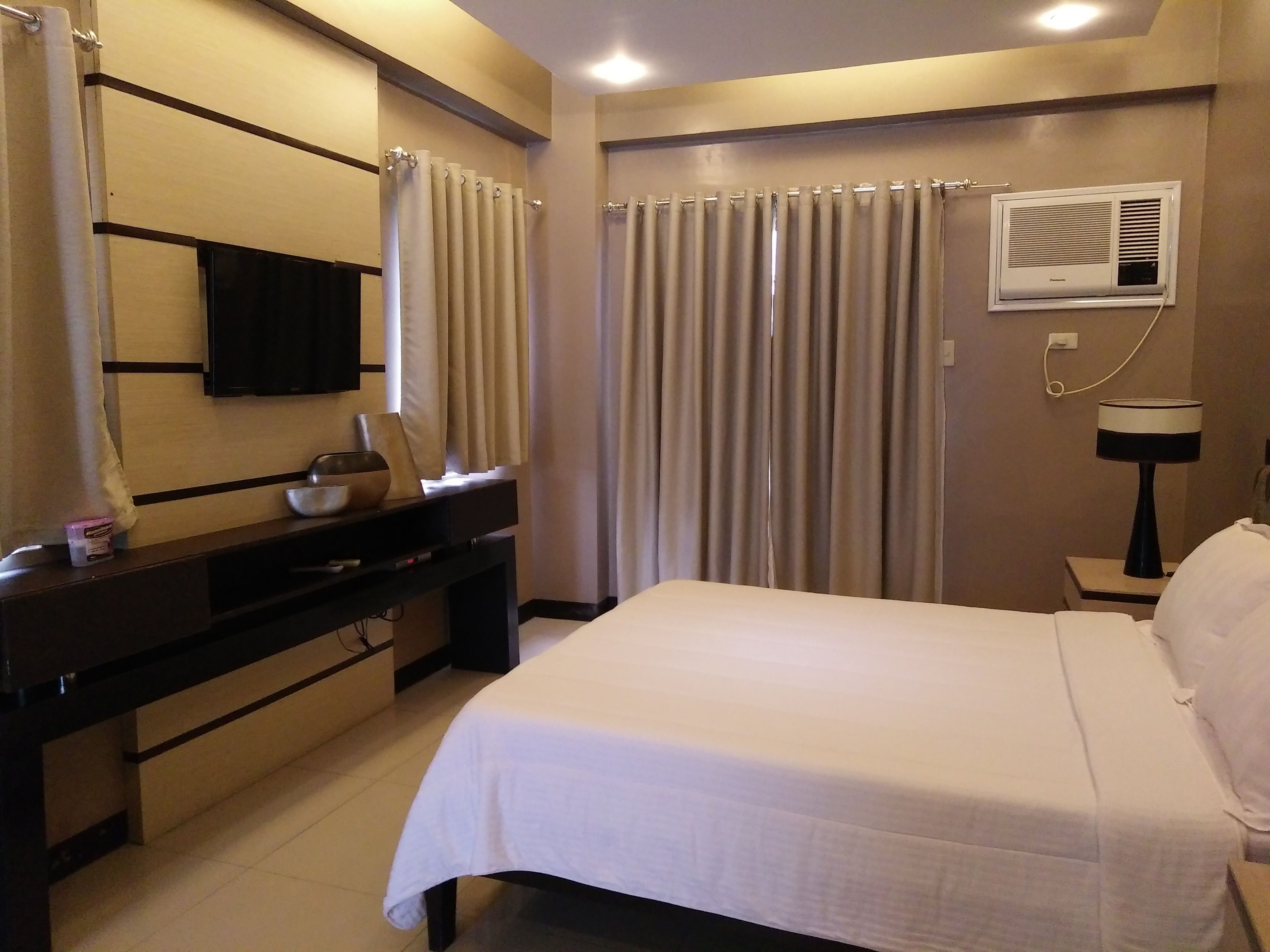 Habitación de hotel con cama, escritorio con espejo, televisor, mesita de noche con lámpara y ventana con cortinas.