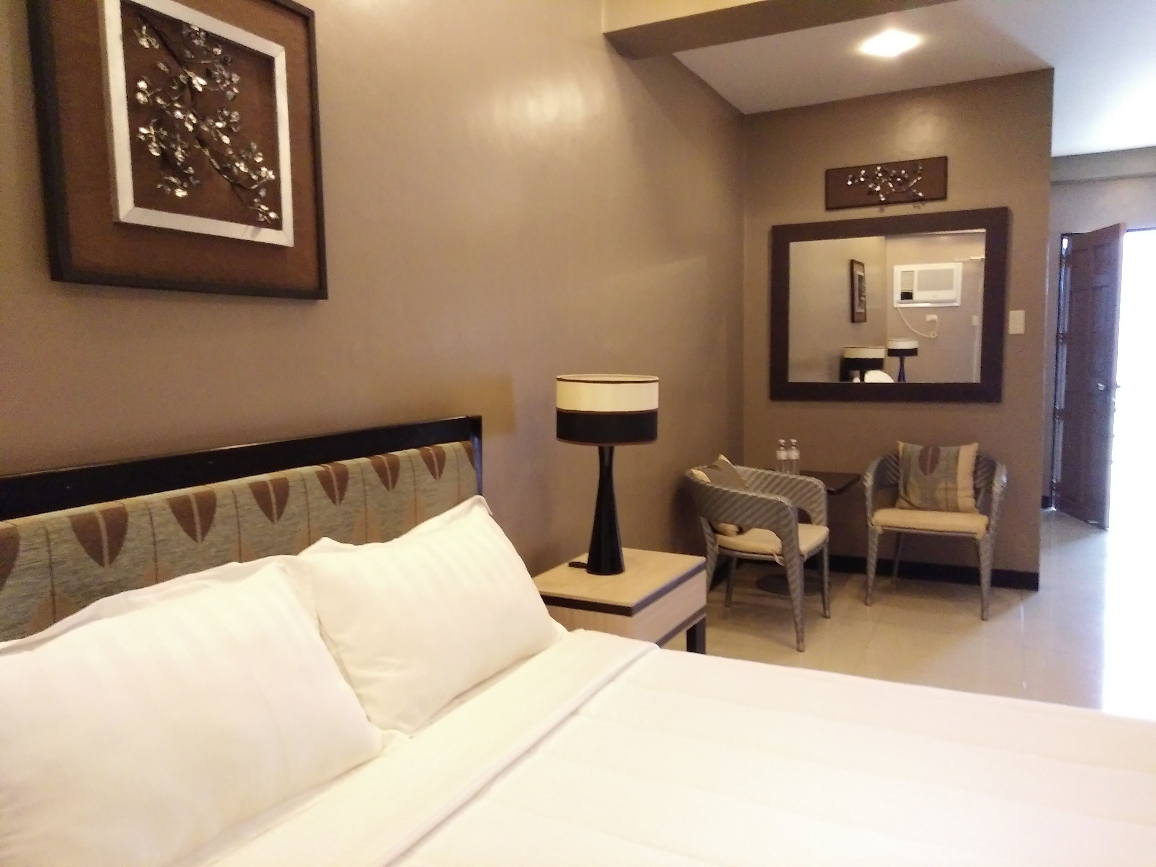 A hotel room with a bed, a desk, a chair, a lamp, a mirror, and a framed artwork on the wall.