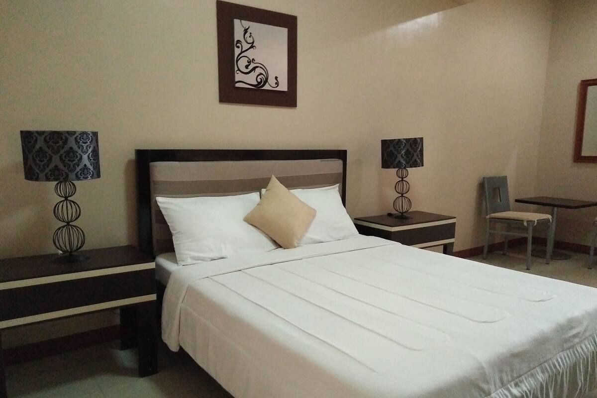 A neatly made bed with white linens, two bedside tables with lamps, a chair, and a framed artwork on the wall.