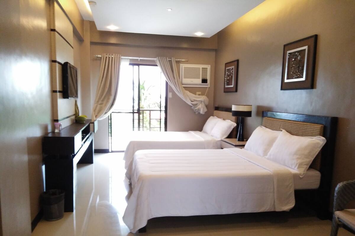 A hotel room with two beds, a TV, a bedside table, a lamp, and a window with curtains.