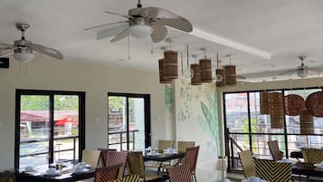 Comedor con mesas listas para el servicio, sillas con tapicería estampada, ventiladores de techo y grandes ventanales con vistas.