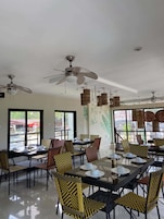 A dining area with tables set for service, chairs with patterned upholstery, ceiling fans, and large windows with views.