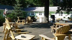 An outdoor seating area with wooden Adirondack chairs and tables, surrounded by trees and a building in the background. - The Pines Motorlodge (Douglas)