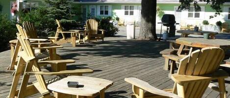 An outdoor seating area with wooden Adirondack chairs and tables, surrounded by trees and a building in the background.
