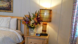 A hotel room with a bed, a nightstand with a lamp, a wicker chair, a table with a wine bottle, glasses, and flowers. - The Pines Motorlodge (Douglas)