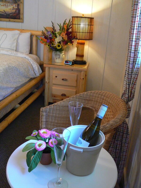 Habitación de hotel con una cama, una mesita de noche con lámpara, una silla de mimbre, una mesa con botella de vino, copas y flores.