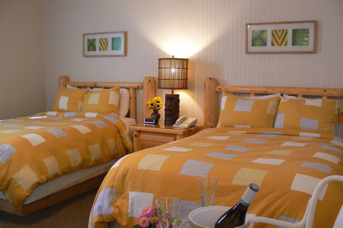 A hotel room with two beds, a nightstand with a lamp, a phone, and a table with a bottle and glasses.