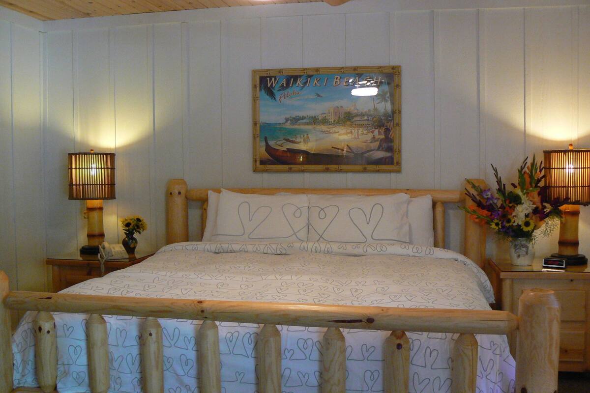 A wooden-framed bed with heart-patterned bedding, two bedside lamps, a wall-mounted painting of Waikiki Beach, and a small vase with flowers.