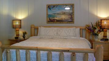 A wooden-framed bed with heart-patterned bedding, two bedside lamps, a wall-mounted painting of Waikiki Beach, and a small vase with flowers.