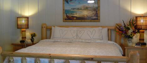 Ein Bett mit Holzrahmen, Bettwäsche mit Herzmuster, zwei Nachttischlampen, ein an der Wand hängendes Bild von Waikiki Beach und eine kleine Vase mit Blumen.