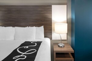 Una cama bien hecha con sábanas blancas y una colcha a rayas negras y blancas, una mesita de noche de madera con una lámpara, y una pared con textura.