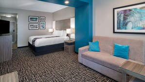 A hotel room with a bed, a sofa, a coffee table, and a TV. There are framed pictures on the wall and a painting above the sofa. - La Quinta Inn & Suites by Wyndham Forsyth (Forsyth)