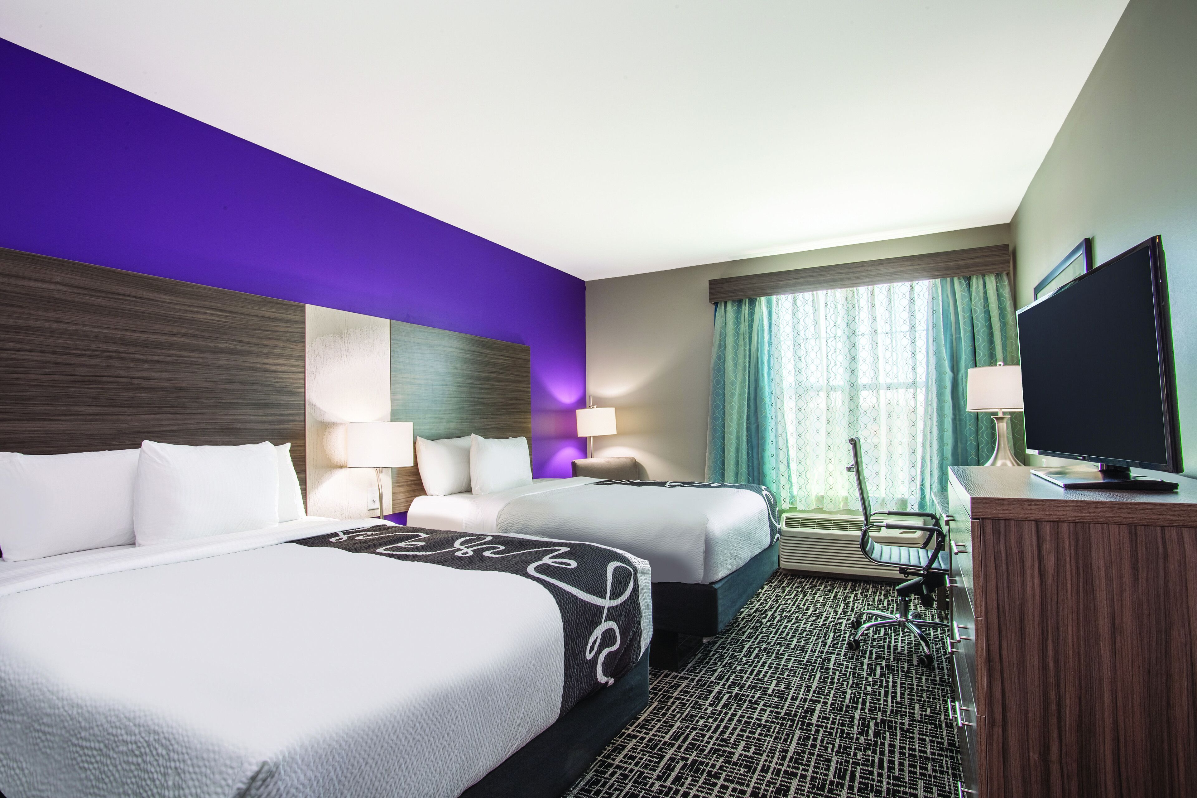 A hotel room with two beds, a desk, a television, and a window with curtains.