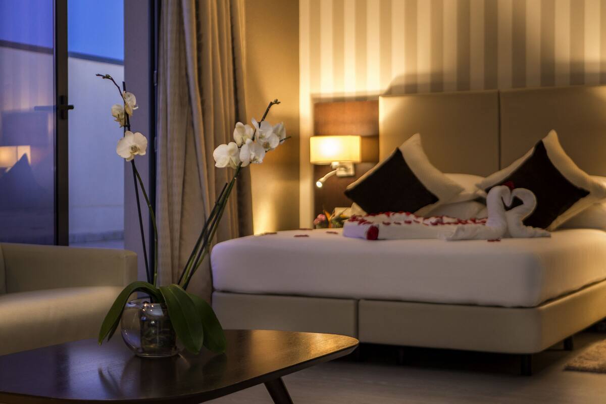 A hotel room with a bed, a bedside table, a lamp, and a vase with flowers.