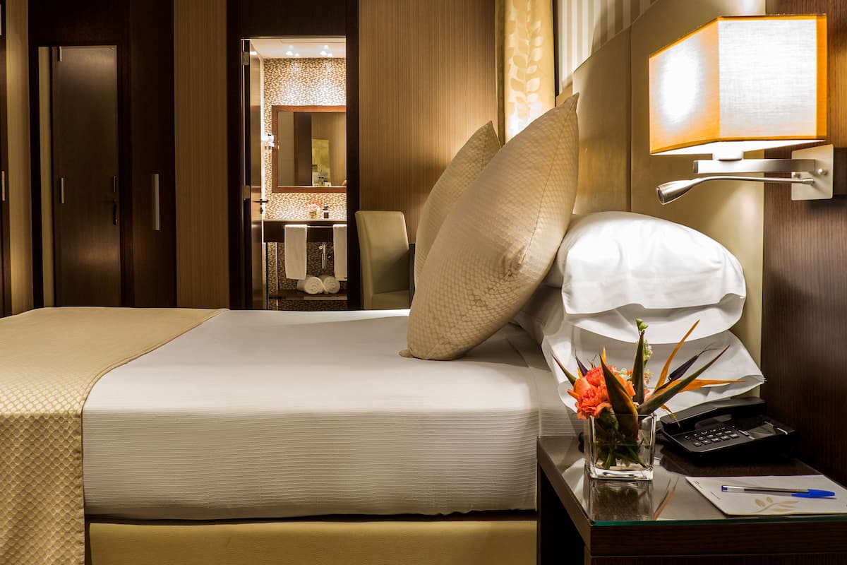 A hotel room with a neatly made bed, a bedside table with a lamp, a telephone, and a vase with flowers.