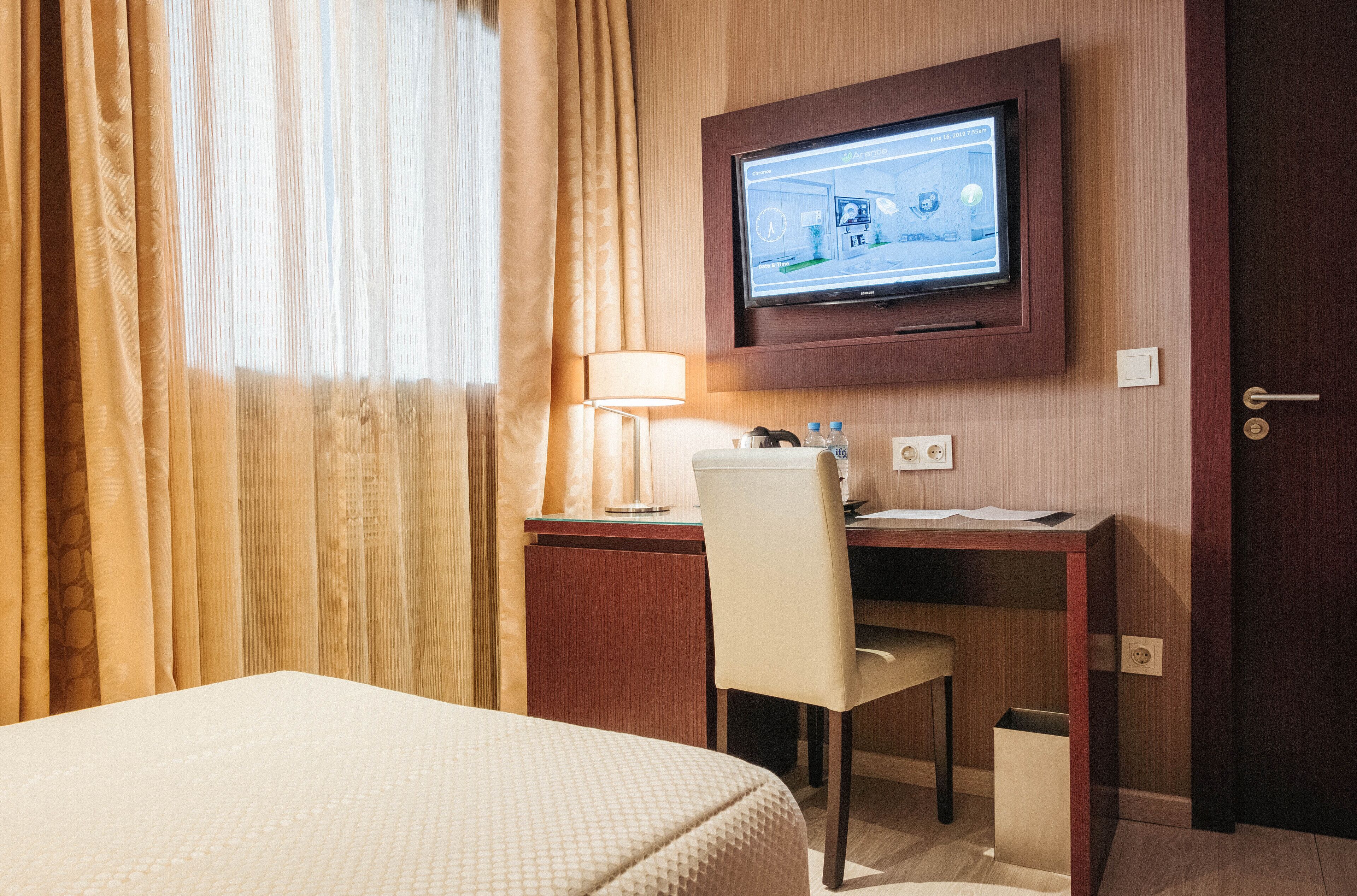 Habitación de hotel con cama, escritorio y silla, televisión montada en la pared y ventana con cortinas.
