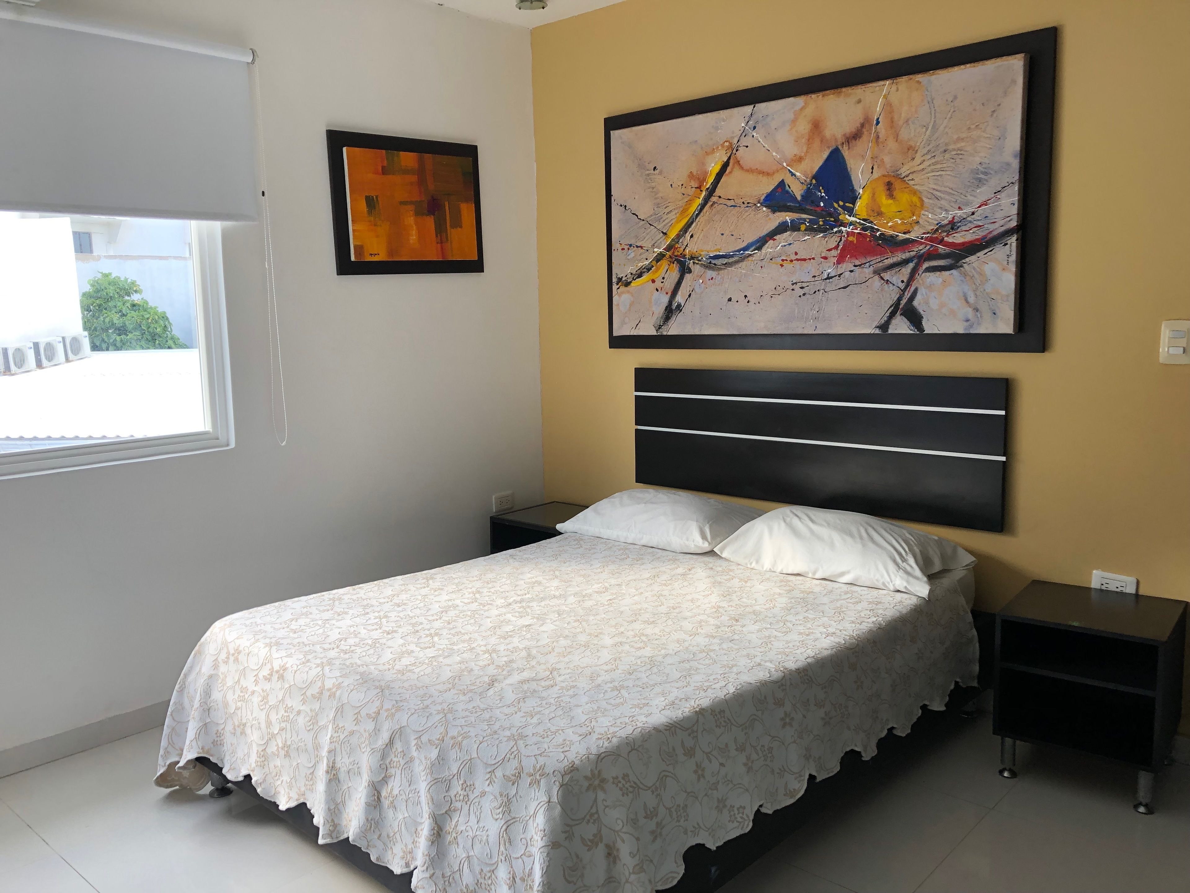 A bedroom with a bed, two nightstands, a window, and two paintings on the wall.