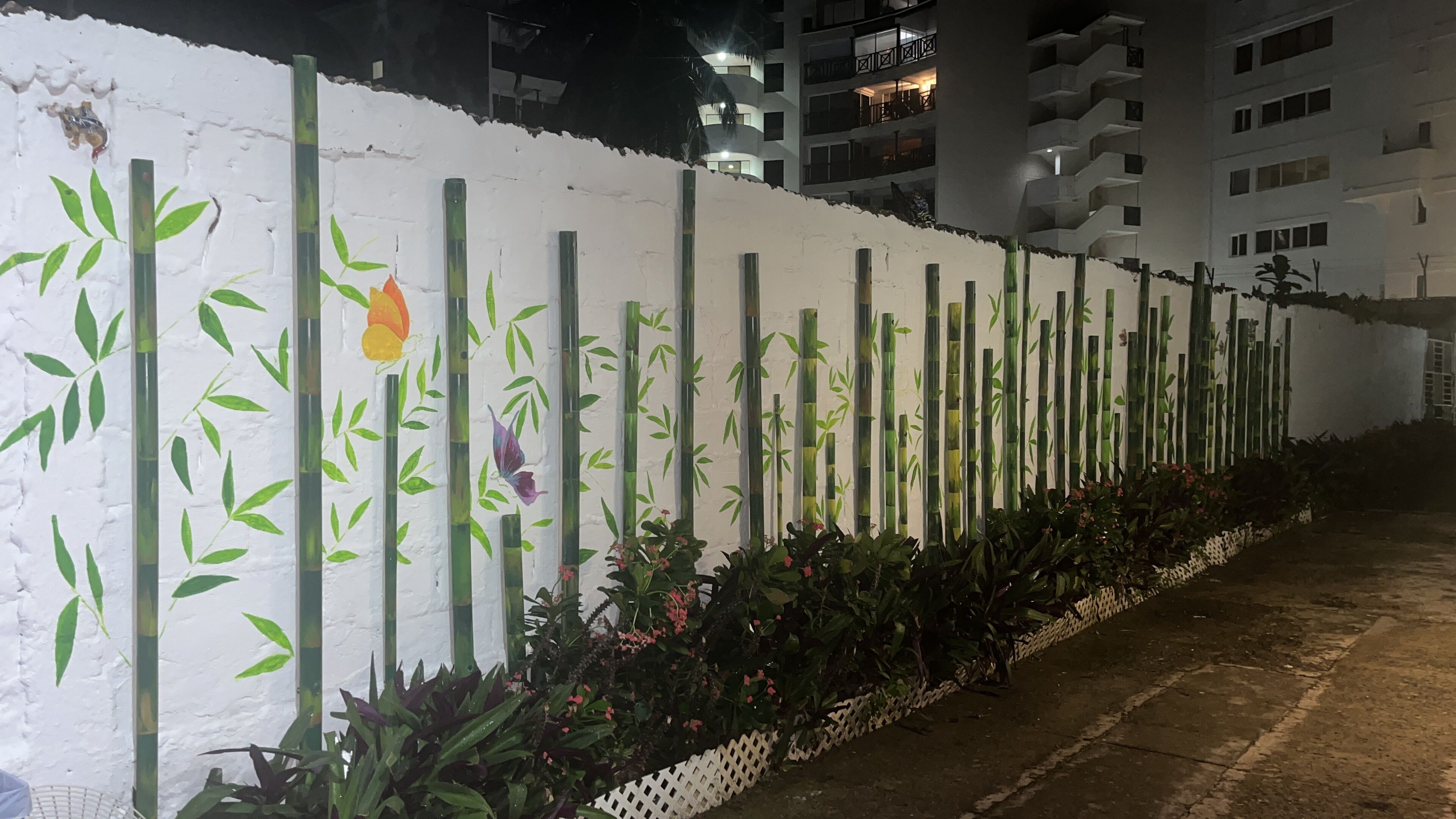 Pared blanca con patrones verticales de bambú y hojas, una valla de piquetes blancos y una fila de plantas florecientes.