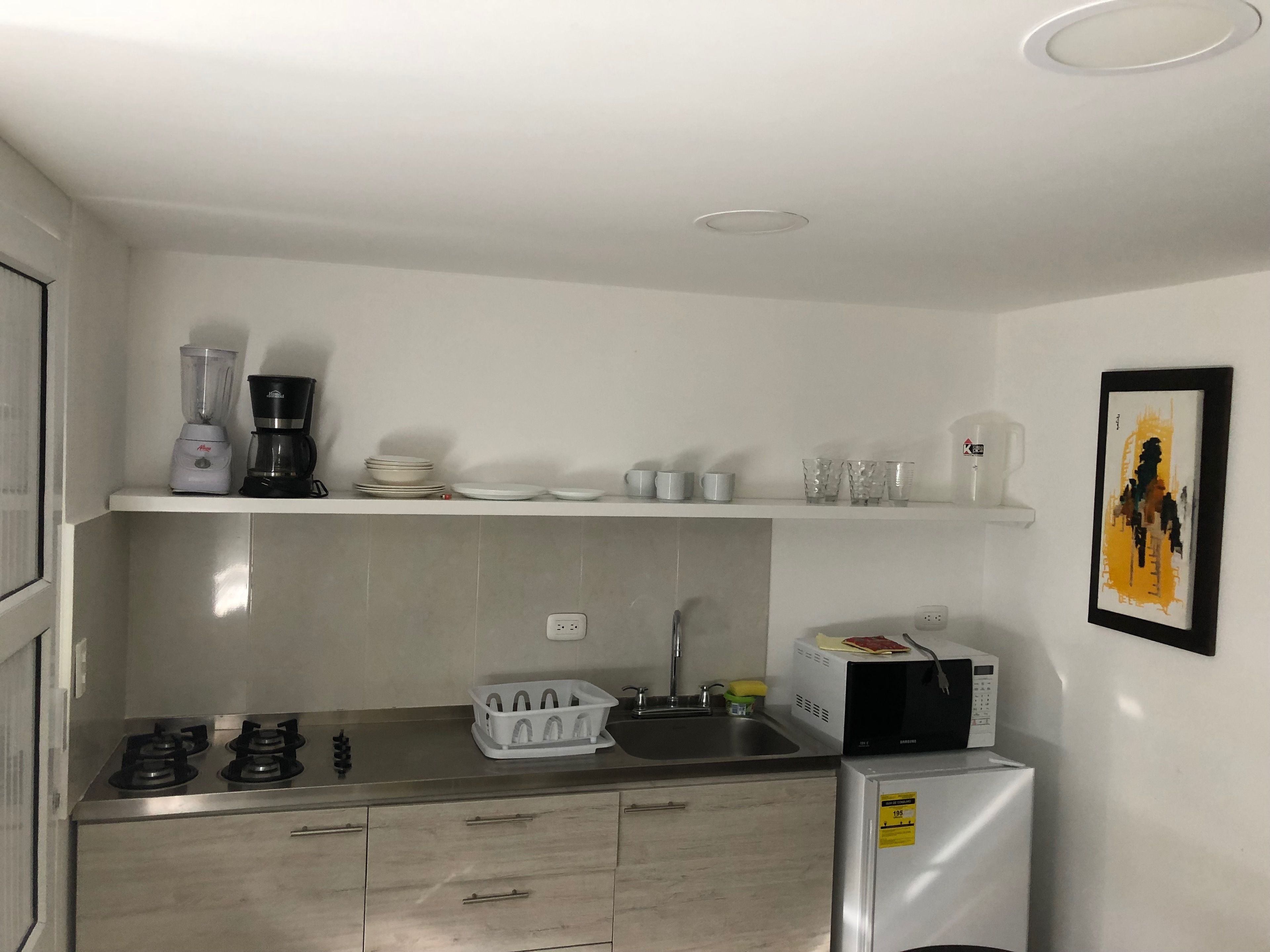 A compact kitchen with a gas stove, a microwave, a refrigerator, and a sink. There are shelves above the counter holding dishes and glasses.