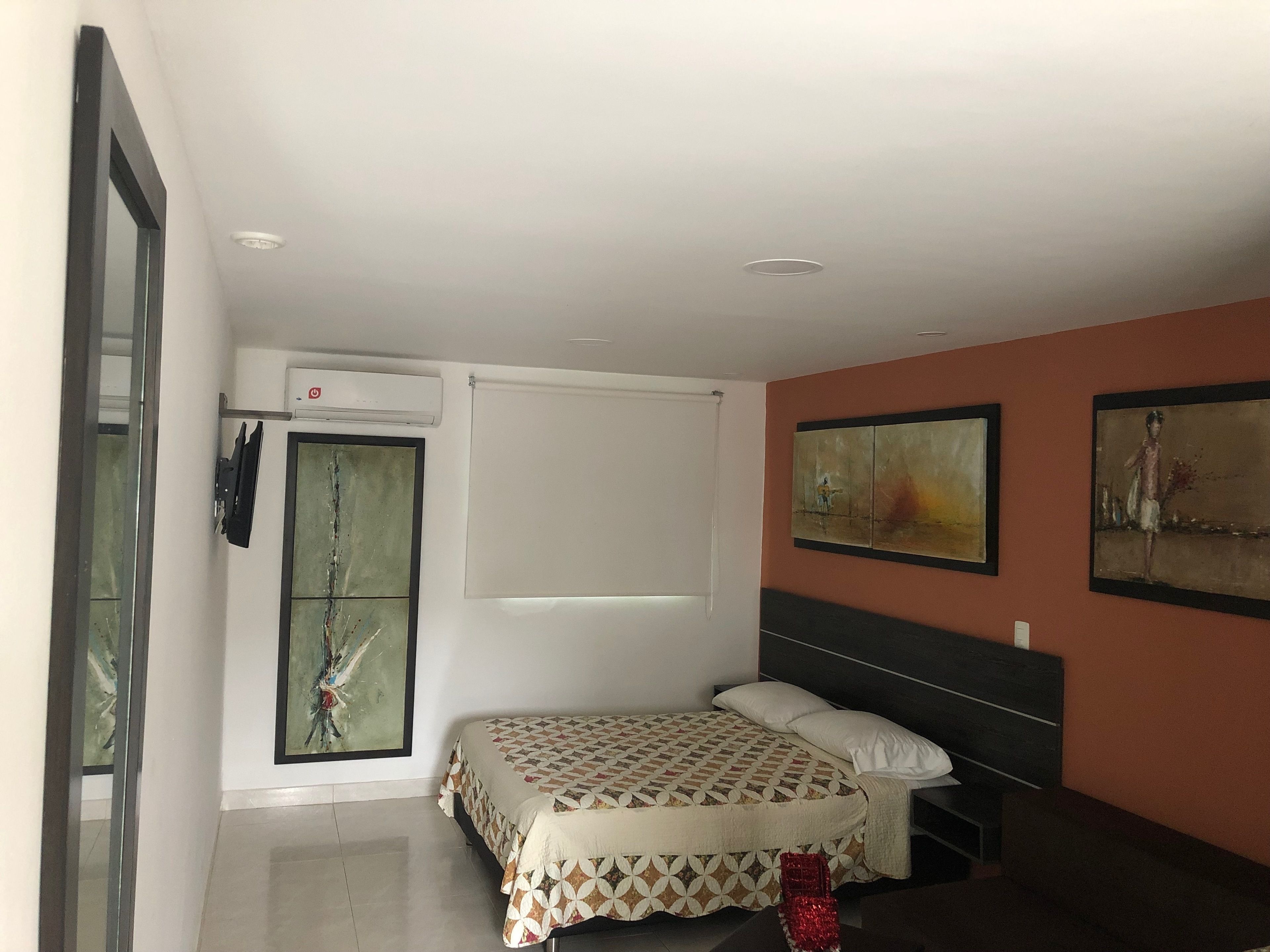 Une chambre à coucher avec un lit, une fenêtre avec des stores, un climatiseur fixé au mur et deux tableaux abstraits.