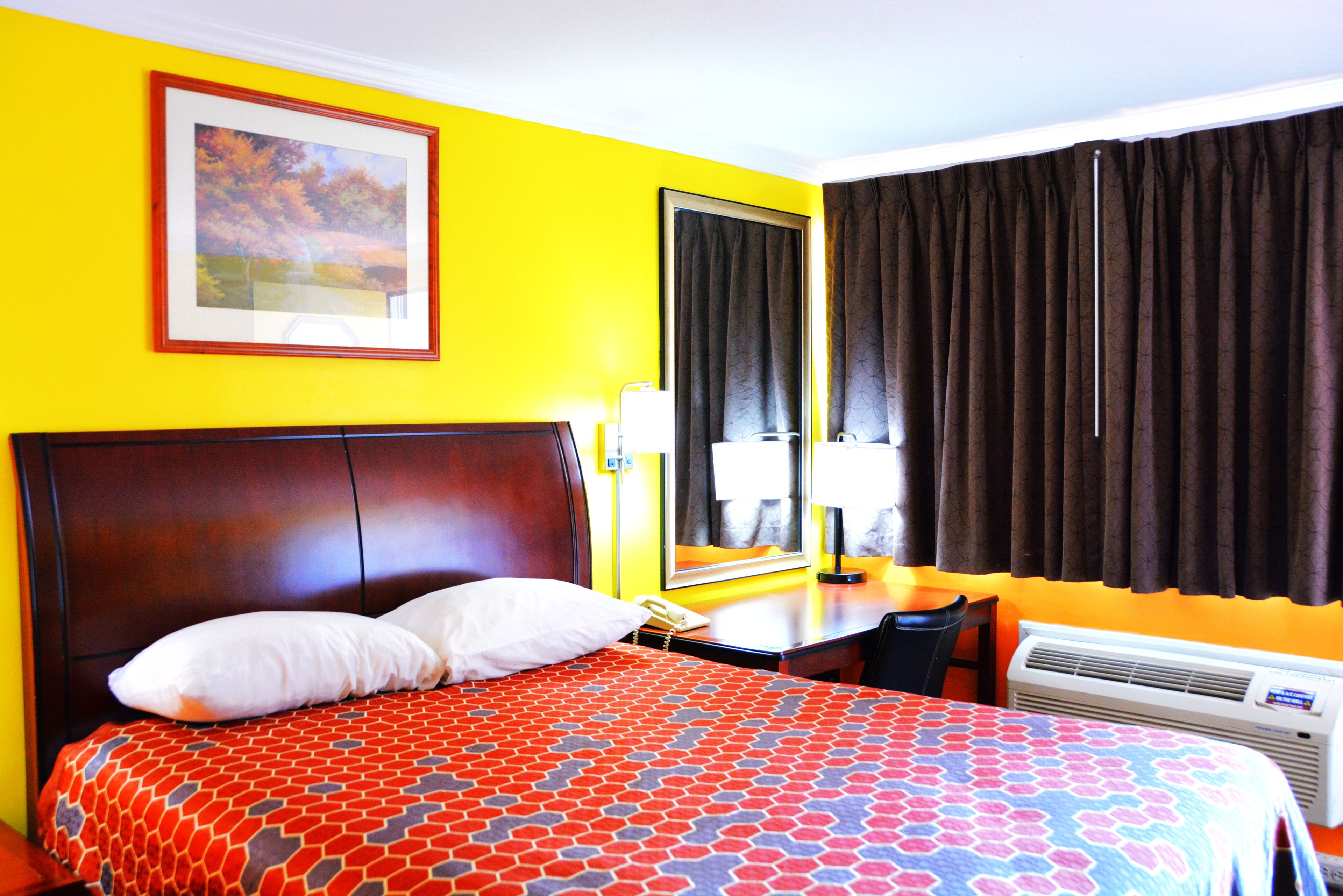 A hotel room with a large bed, a bedside table with a lamp, a telephone, and an air conditioning unit.