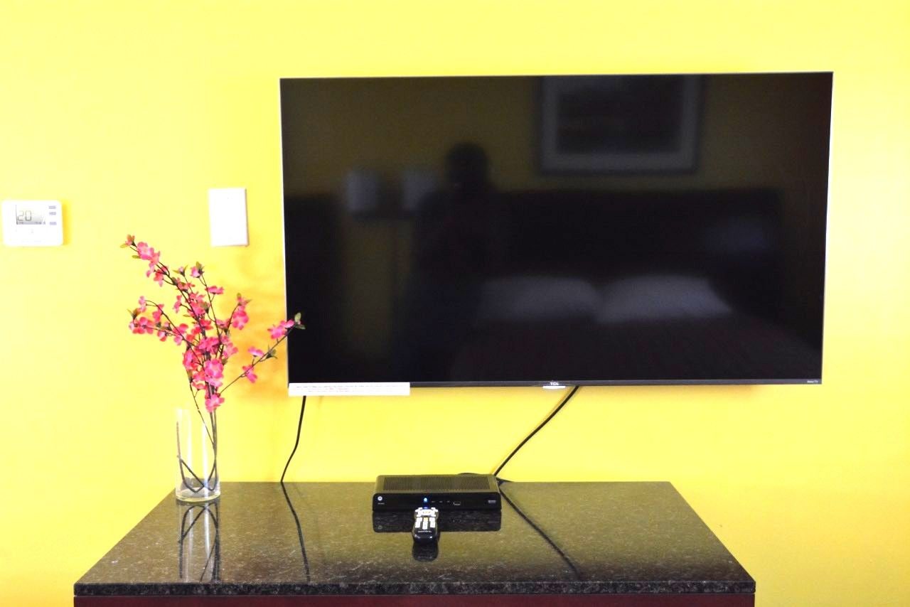 Un televisor de pantalla plana montado en la pared, un jarrón pequeño con flores rosas sobre una mesa y un termostato digital.