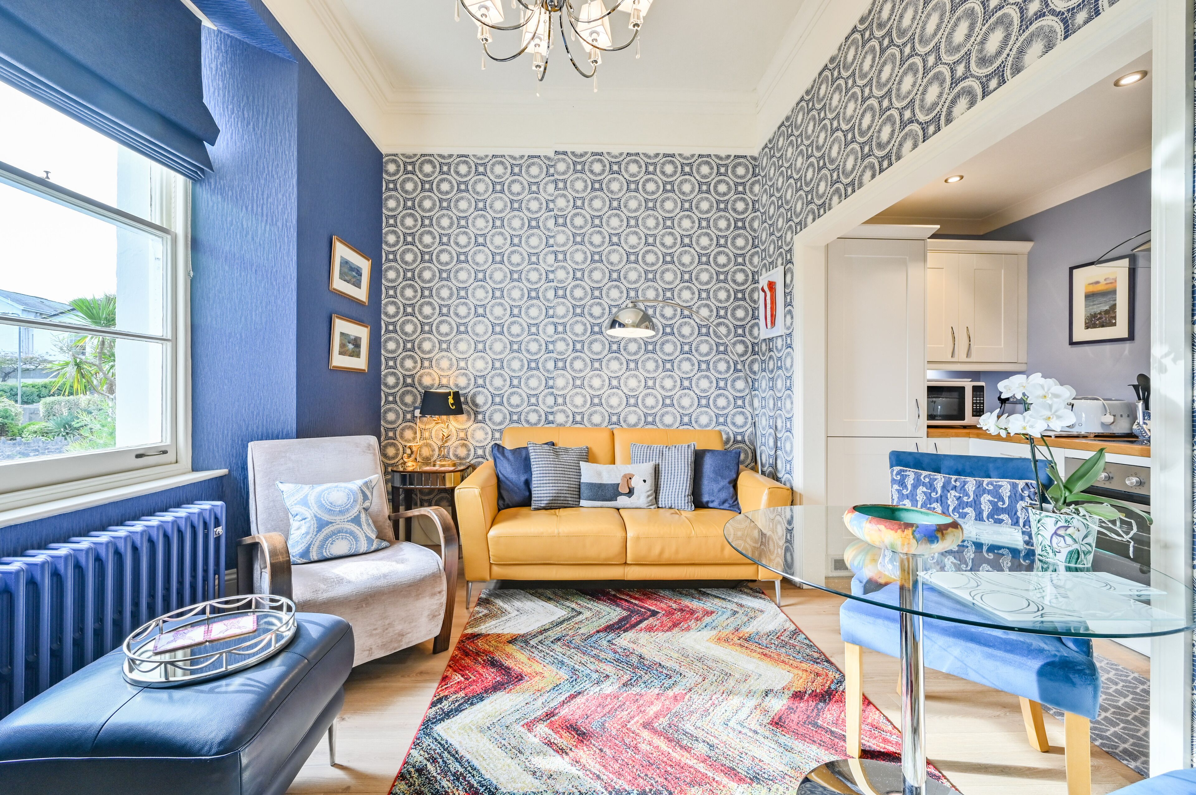 A modern living room with a yellow sofa, blue and white patterned wallpaper, a glass coffee table, and a blue sofa with patterned cushions.