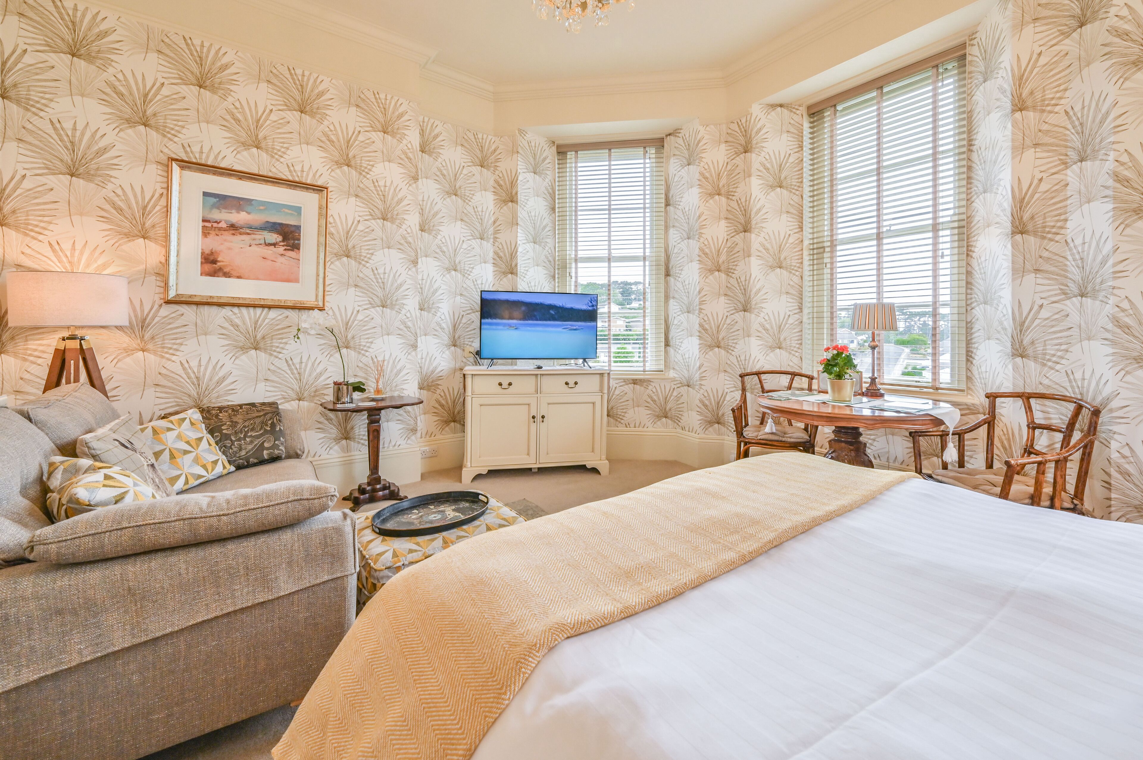 A hotel room with a bed, a sofa, a TV, a small table with a lamp, and a window with blinds.