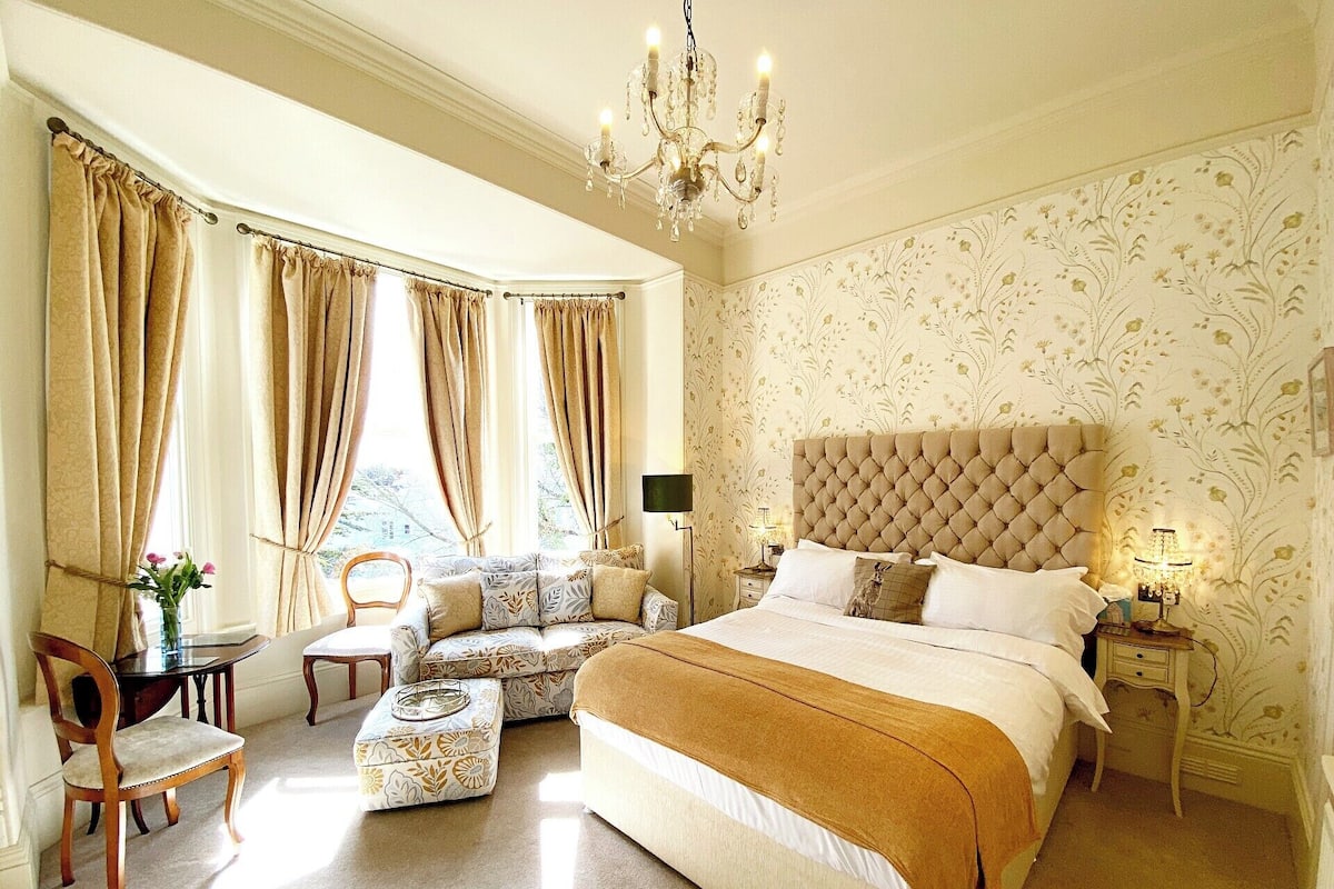 A spacious bedroom with a large bed, a sitting area with a sofa and chairs, and a chandelier.