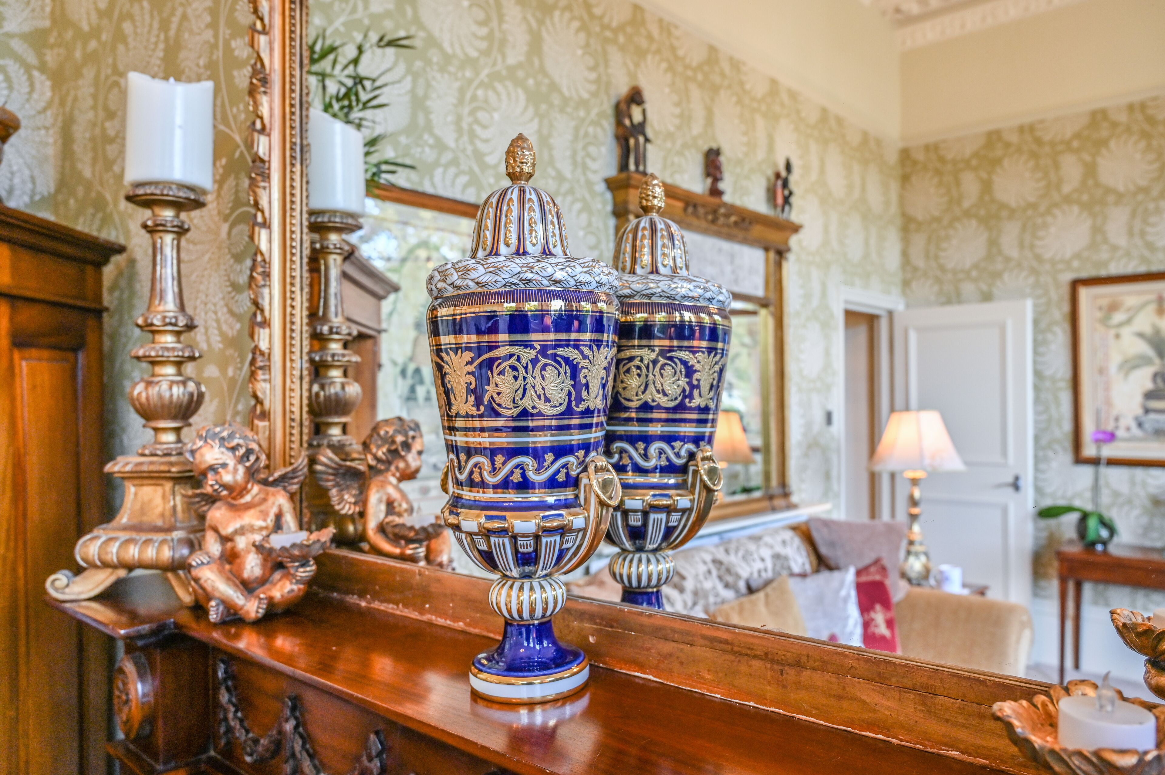 Ornate blue and gold vases on a wooden shelf, a mirror reflecting a room with patterned wallpaper and a lamp.