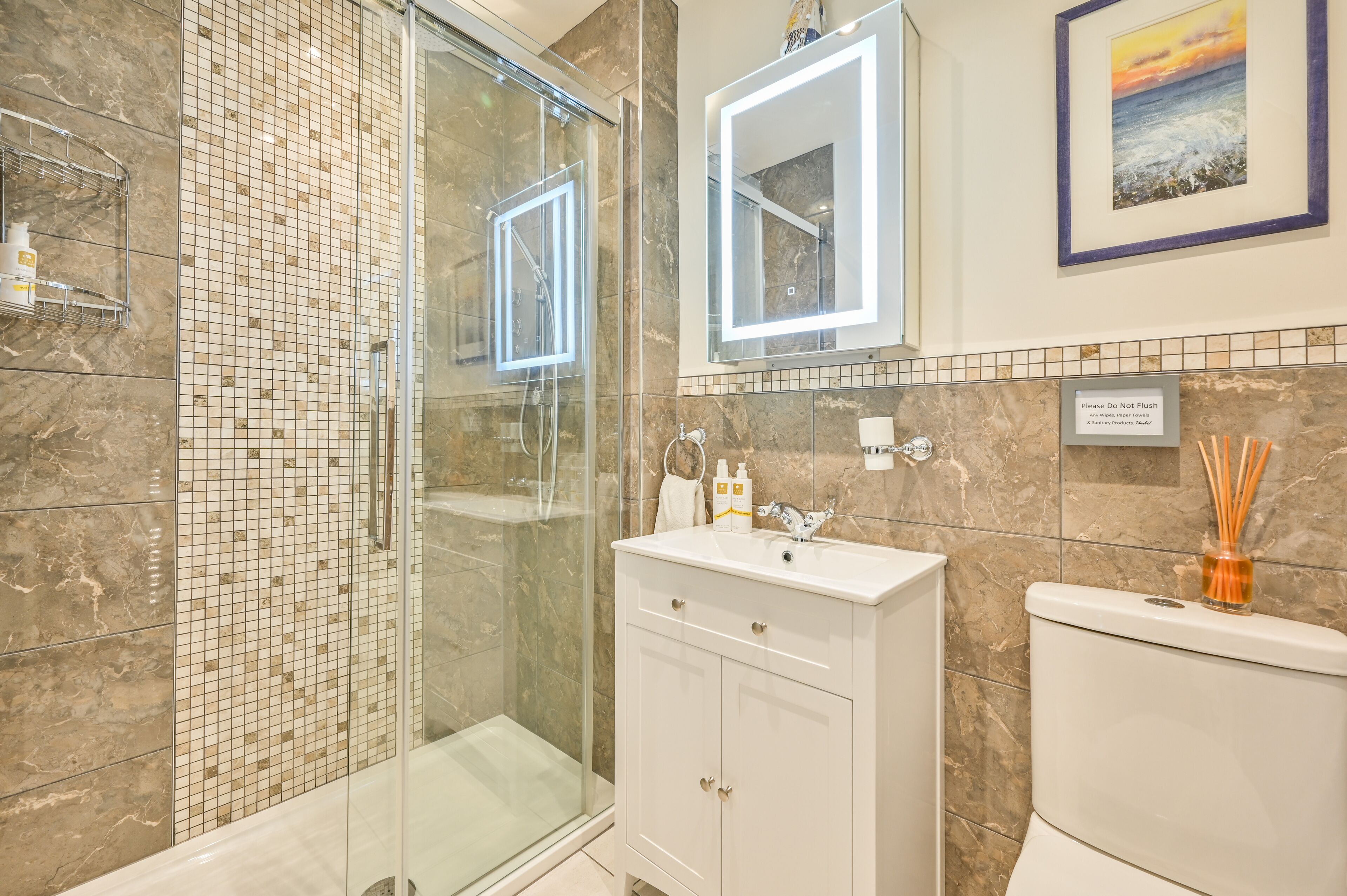 Un baño moderno con ducha envuelta en vidrio, un tocador blanco con espejo y un inodoro decorado con un jarrón.