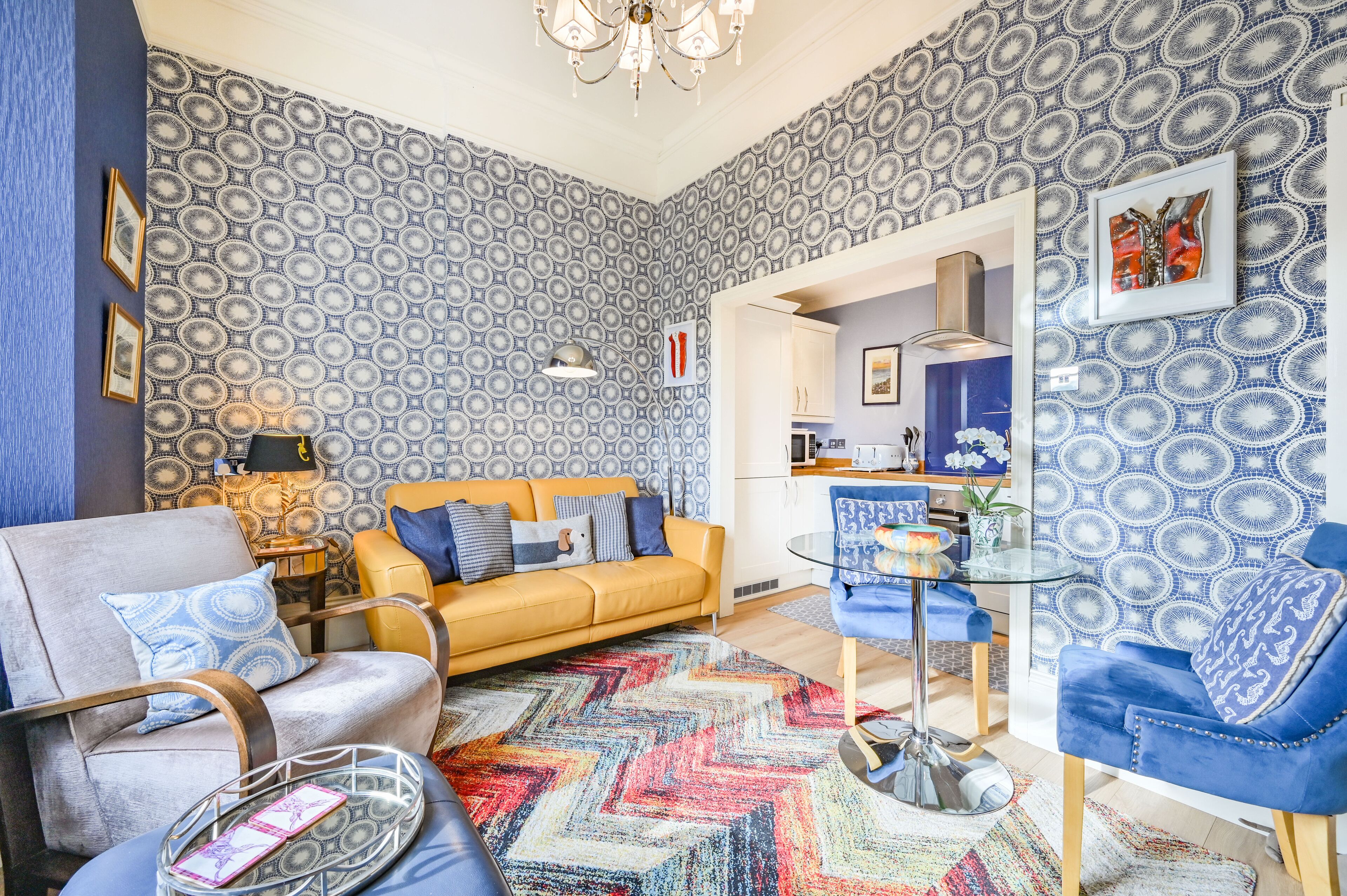 A living room with a patterned wallpaper, a yellow sofa, a blue armchair, a glass coffee table, and a colorful rug.
