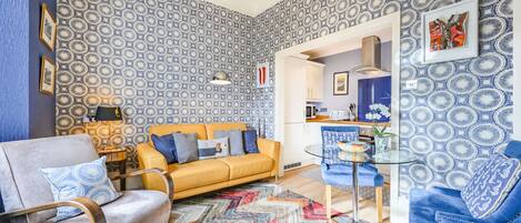 A living room with a patterned wallpaper, a yellow sofa, a blue armchair, a glass coffee table, and a colorful rug.