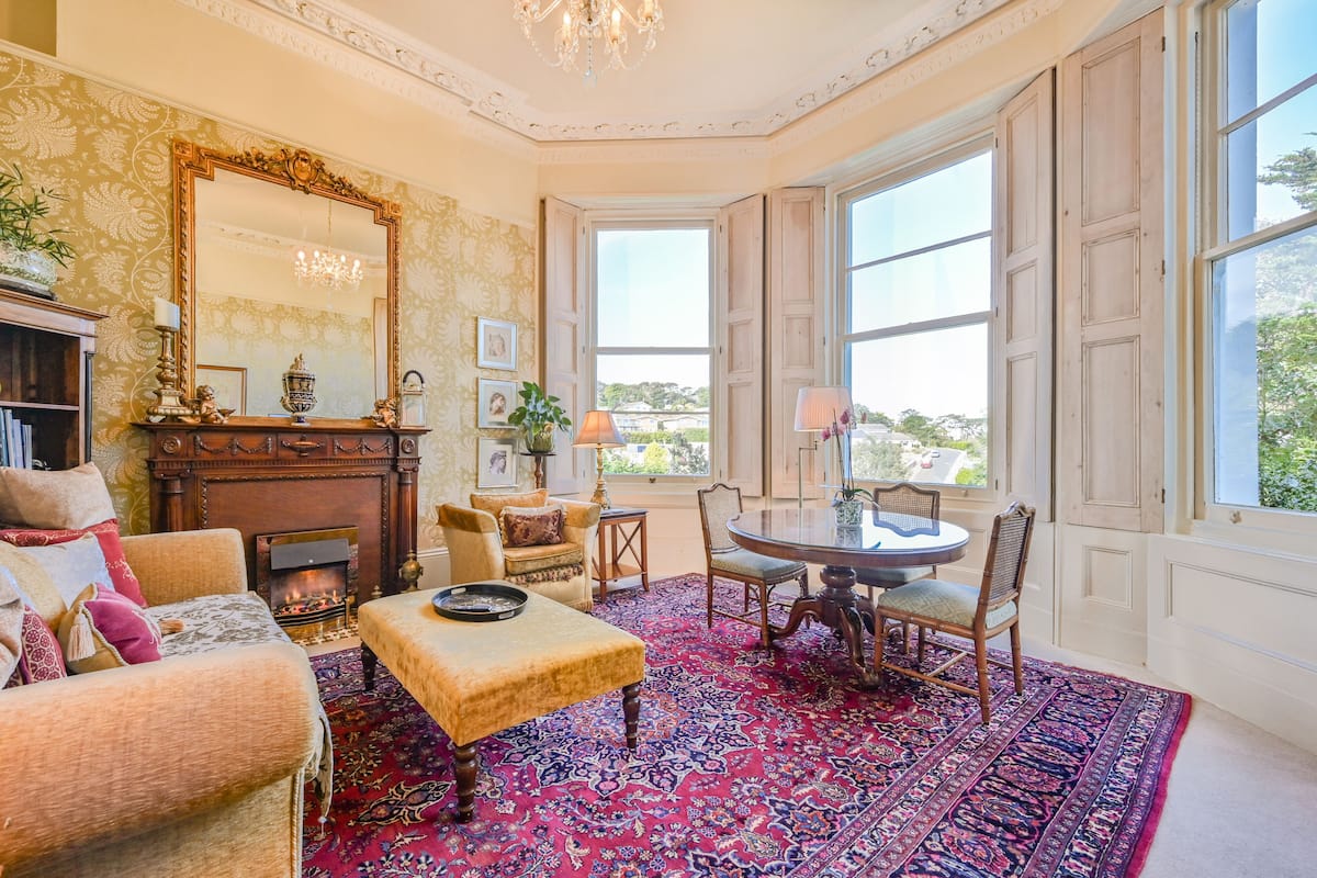 A traditional room with a fireplace, a large mirror, a dining table with chairs, and a view through a bay window.