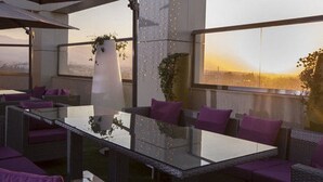An outdoor seating area with a glass-top table and wicker furniture, surrounded by large windows offering a view of the cityscape. - KT Hotels (Ouled Yaich)