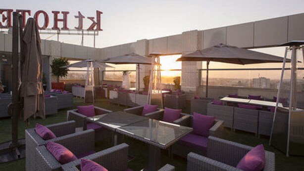 An outdoor seating area with wicker furniture and umbrellas, a large sign in the background, and a city view. - KT Hotels (Ouled Yaich)