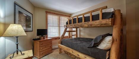 A bunk bed with a ladder, a desk with a lamp, a television, and a window with blinds.