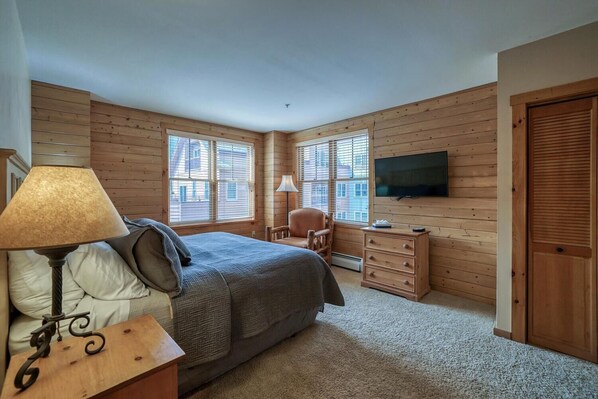 Un dormitorio con paredes de madera, una cama, un escritorio, una silla, un televisor y una ventana.