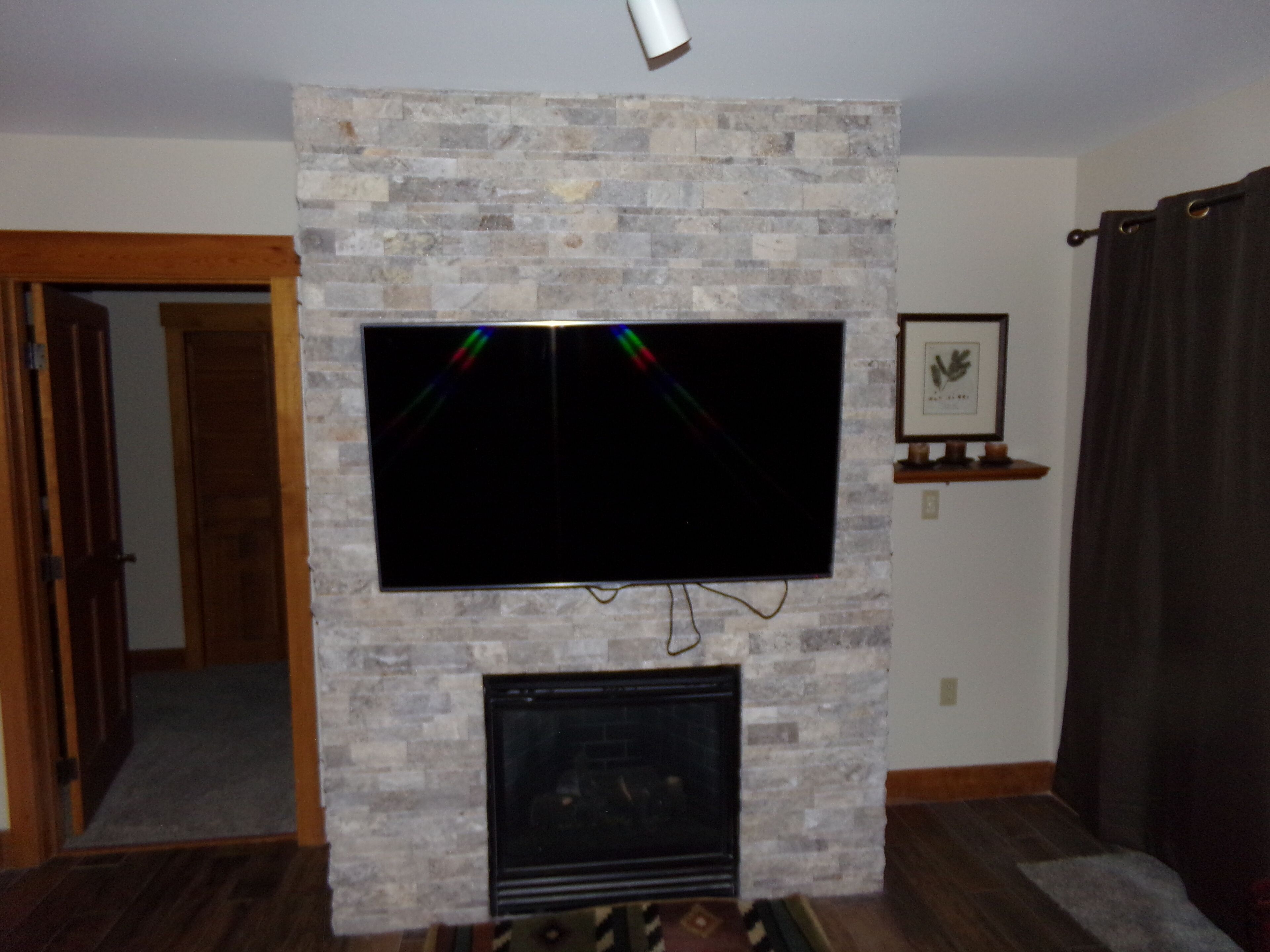 A living room with a flat-screen TV mounted on a stone wall, a fireplace below, and a door leading to another room.