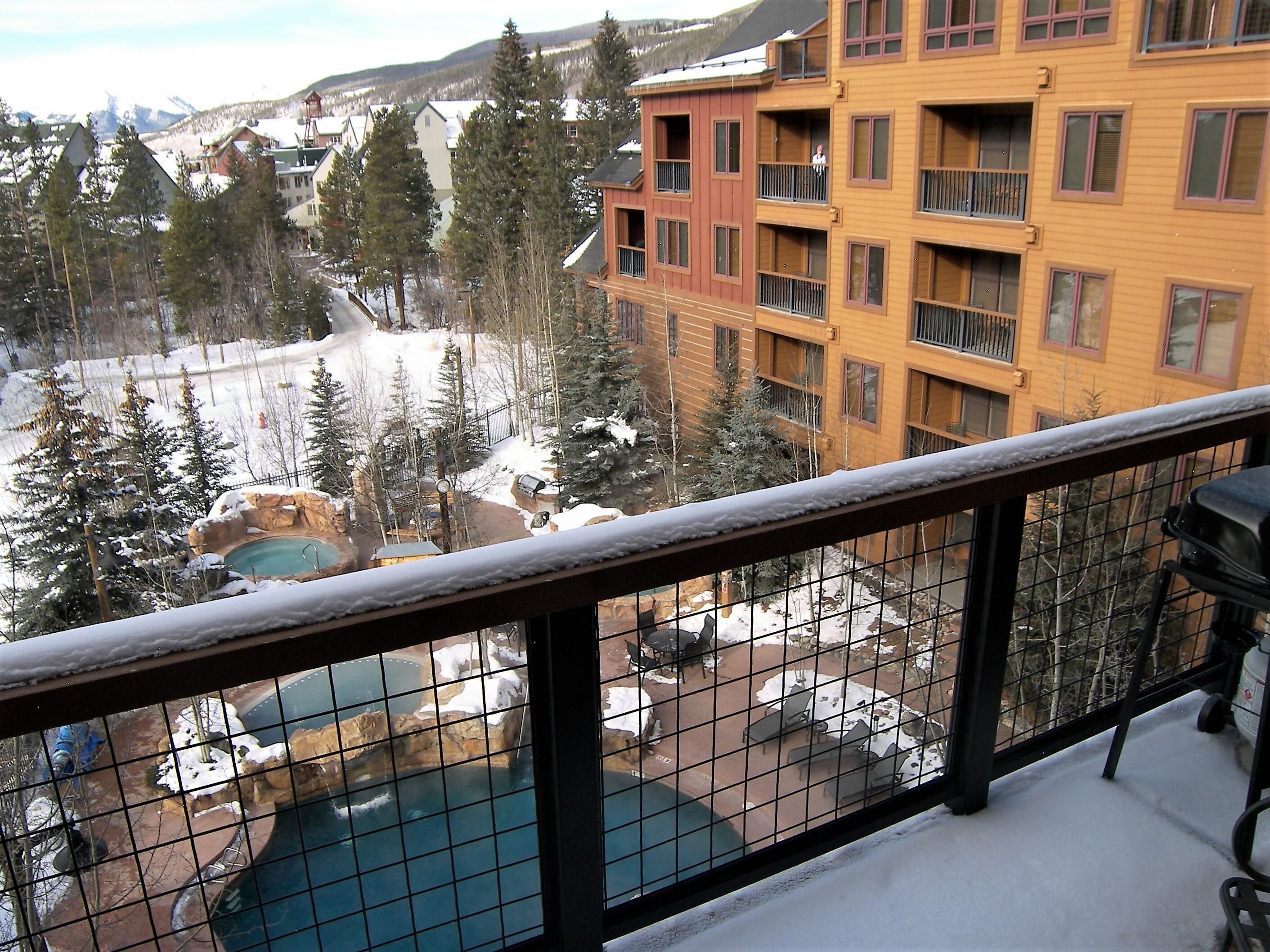 A view from a balcony overlooking a pool area with a snow-covered landscape, a mountain in the background, and a multi-story building with balconies.