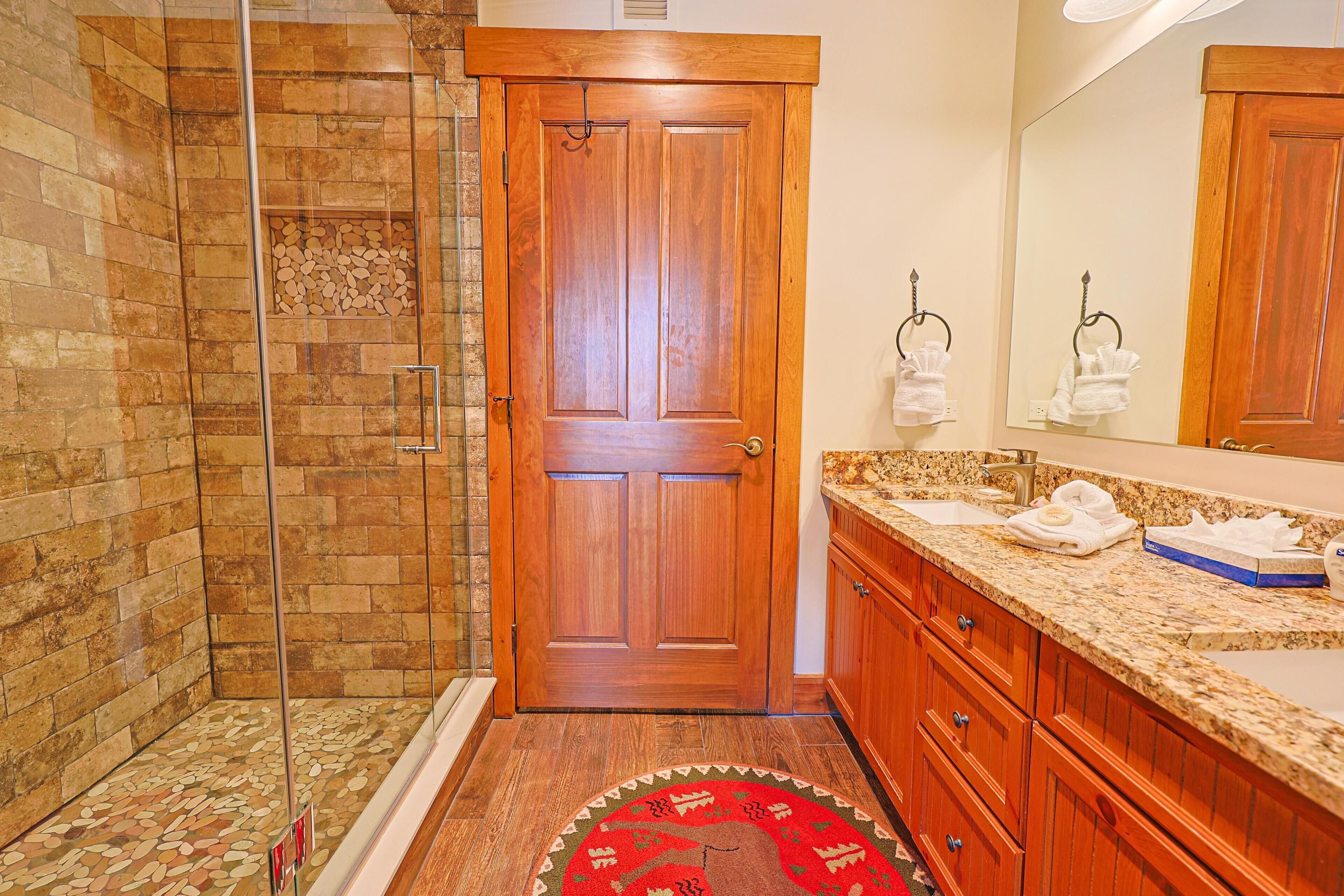 A bathroom with a wooden door, a glass shower enclosure, a granite countertop, and a red patterned rug.